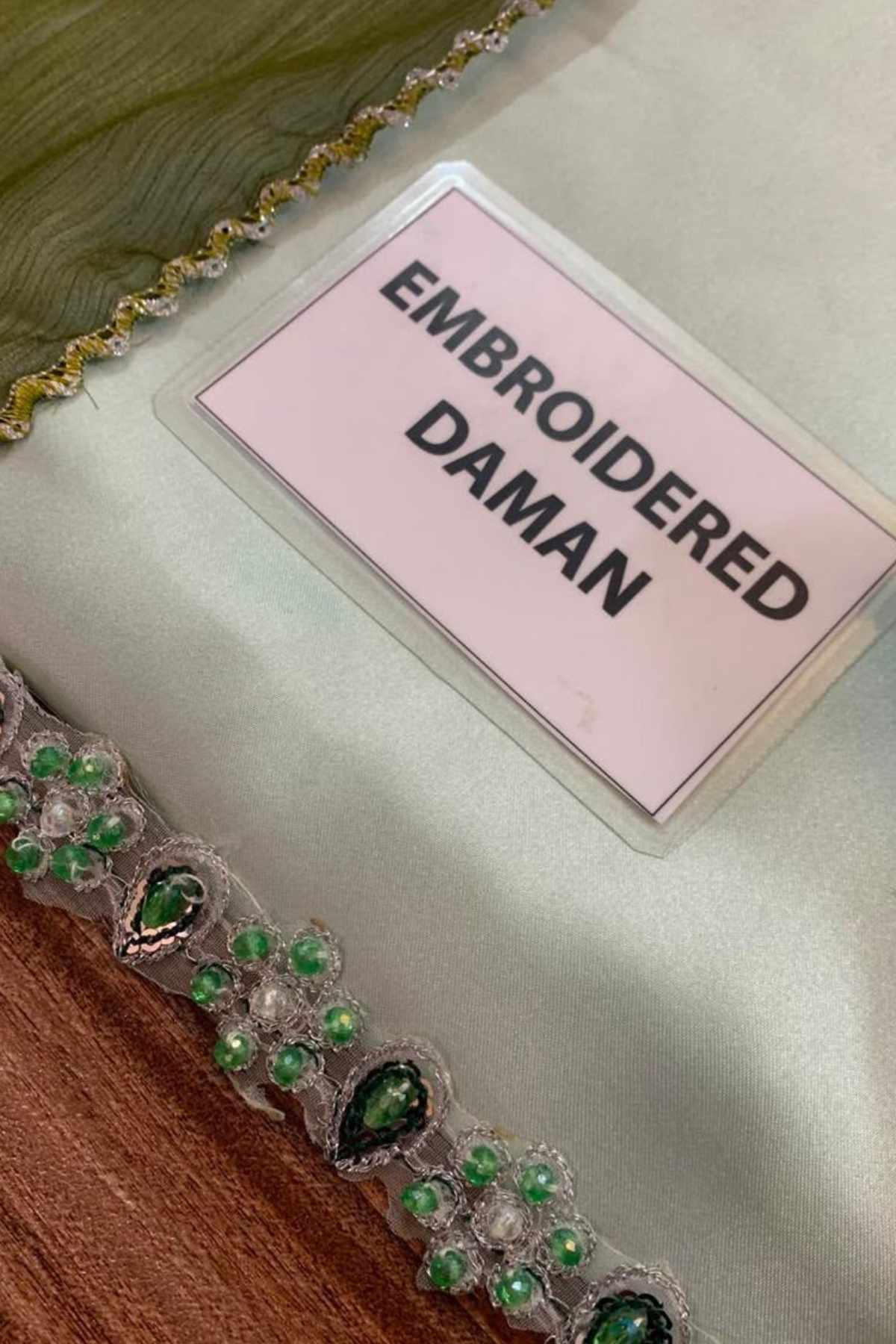 A close-up of an embroidered damask fabric trim with green teardrop-shaped stones and clear rhinestones, featuring a pink label that reads 'EMBROIDERED DAMAN'.