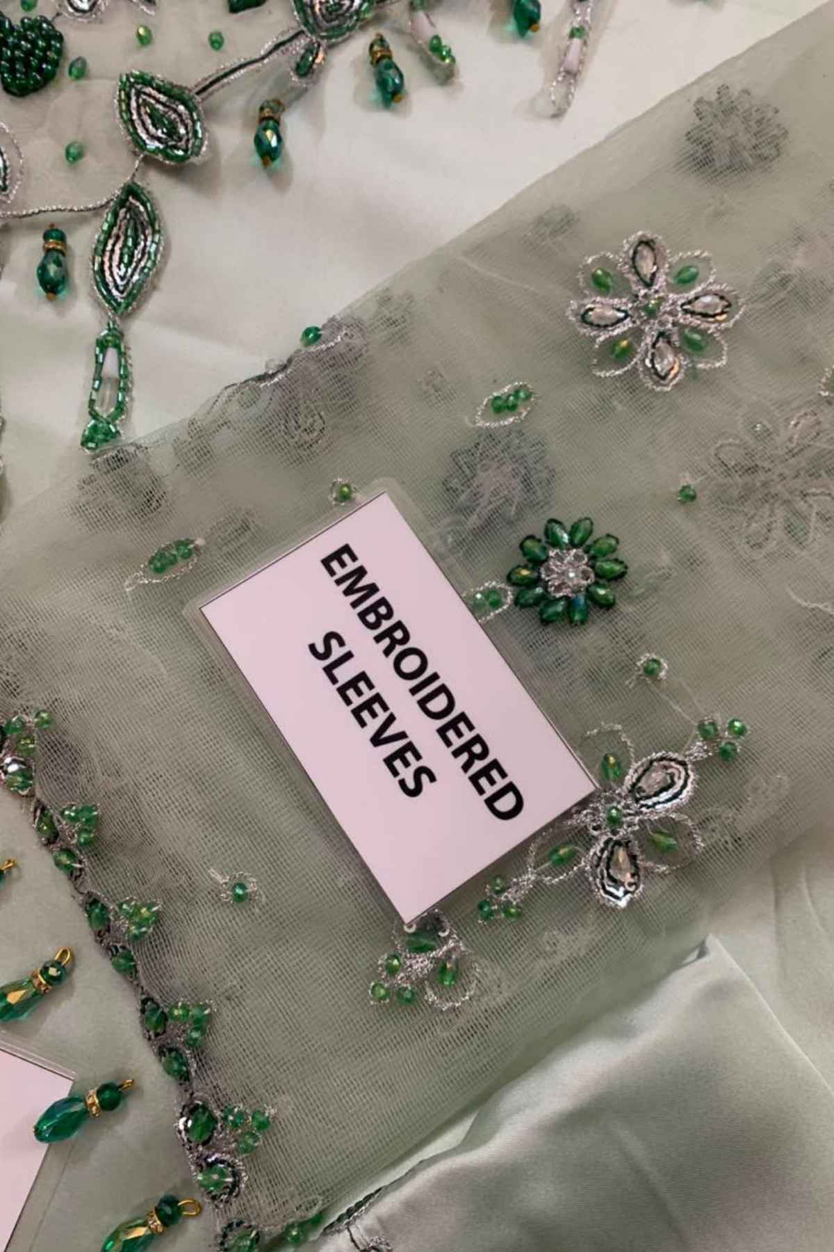A close-up of embroidered tulle sleeves in a pale sage green color, adorned with green and silver beadwork and sequins. A small white label reading 'EMBROIDERED SLEEVES' is placed on the fabric.