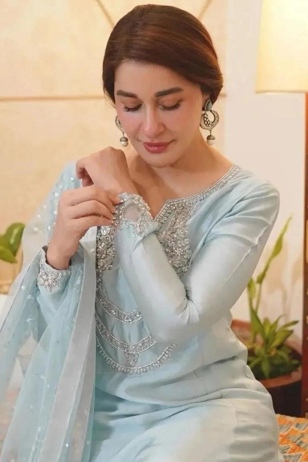A woman wearing a light blue traditional embroidered outfit with silver embellishments, seated indoors with a lamp and plants in the background.
