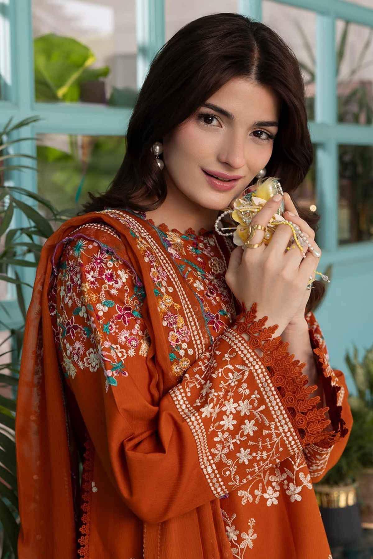 A woman wearing a rust-orange embroidered traditional outfit with floral detailing and a matching dupatta, holding a gold-toned perfume bottle in front of a turquoise window and green plants.