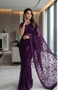 A woman models a deep purple sequined saree with a matching sheer embroidered blouse and a long draped pallu, set in a modern, well-lit interior room.