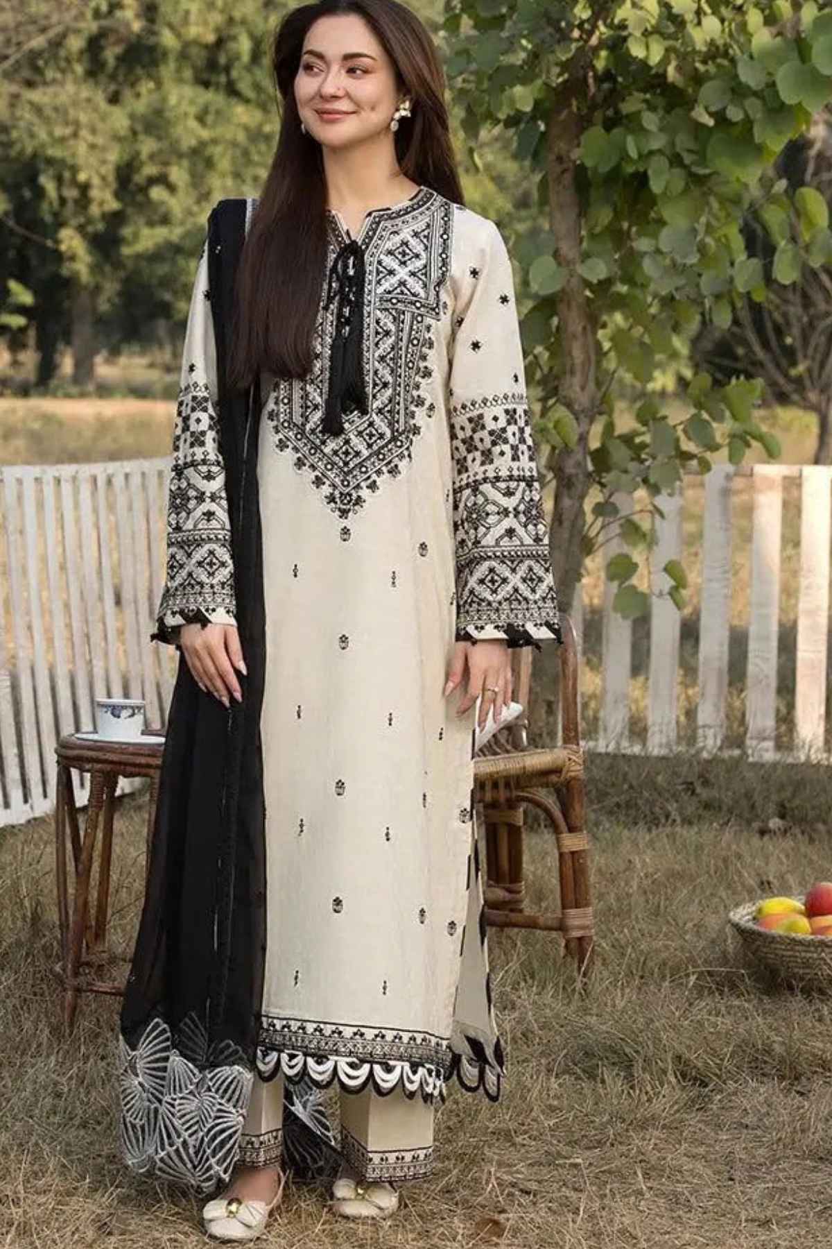 A woman stands outdoors wearing a traditional three-piece salwar kameez suit. The kurta is off-white with black geometric embroidery, paired with matching trousers and a black dupatta featuring a black-and-white patterned border.