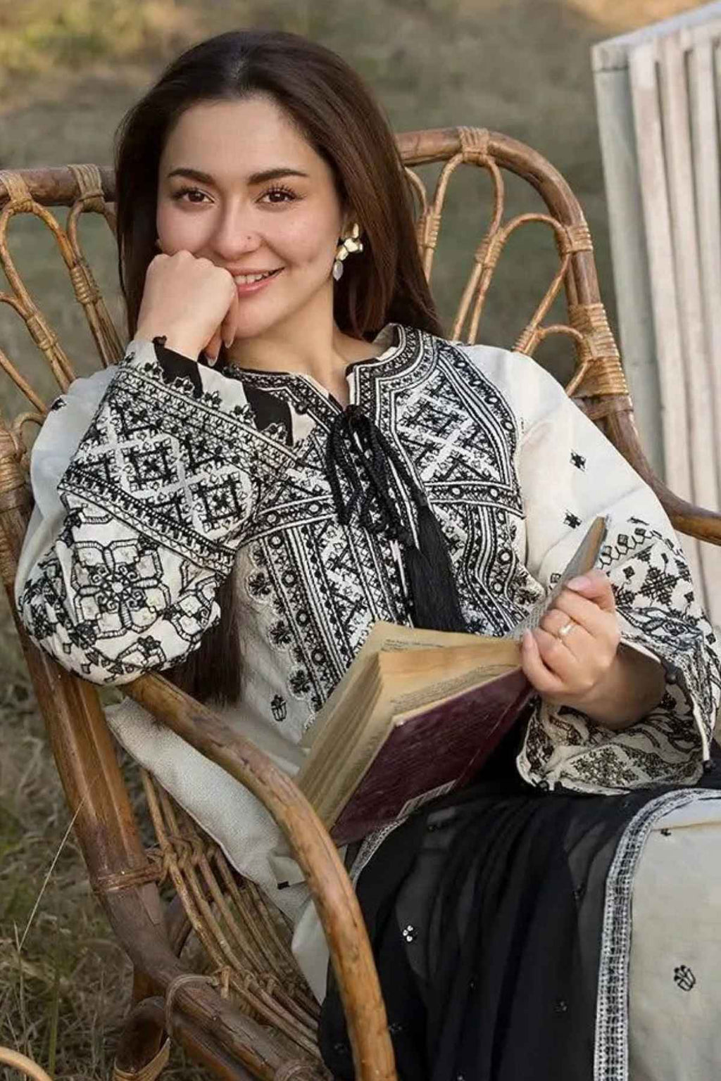 A woman sitting in a rattan chair outdoors, wearing a black and white patterned embroidered top and holding an open book.