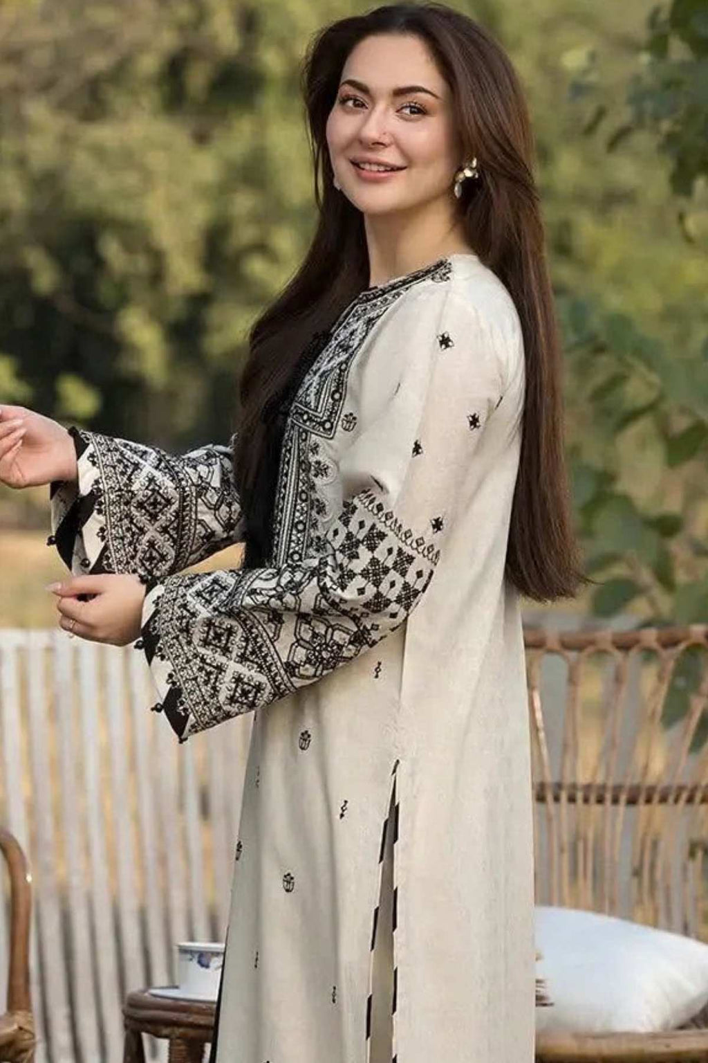 A woman wearing a cream-colored embroidered tunic with black geometric patterns and small metallic embellishments, posed outdoors in front of a softly blurred green background.