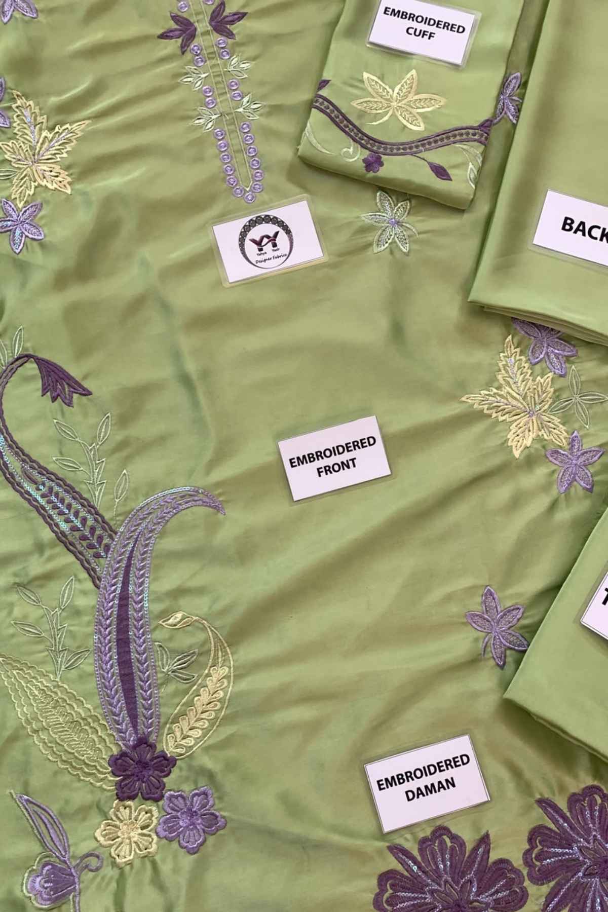 A close-up of a light olive green embroidered fabric, likely a kurta or dress material, showing detailed floral and paisley embroidery in purple, gold, and green. Several white fabric labels are placed on the garment, reading 'EMBROADERED FRONT', 'EMBROADERED DAMAN', 'EMBROADERED CUFF', and 'BACK'.