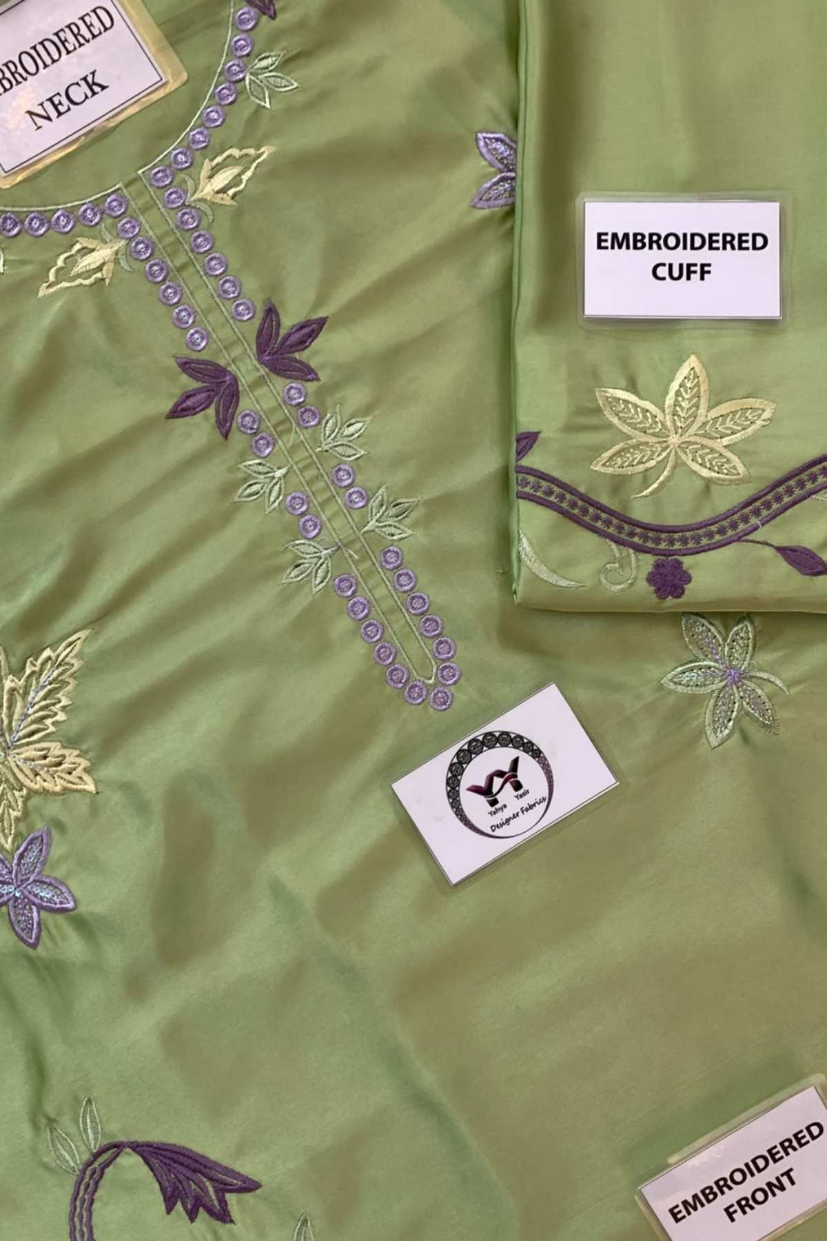 A close-up of an olive green embroidered fabric garment, showing the front bodice with purple and gold floral embroidery, a folded matching cuff with embroidered border, and fabric labels reading 'EMBROIDERED NECK,' 'EMBROIDERED CUFF,' and 'EMBROIDERED FRONT.'.
