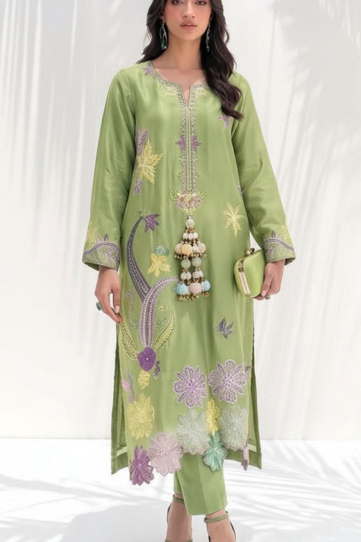 A woman models a light green three-piece salwar kameez suit with floral embroidery in purple, yellow, and white. The set includes a long-sleeved tunic with a split neckline, matching trousers, and a decorative beaded tassel accessory.