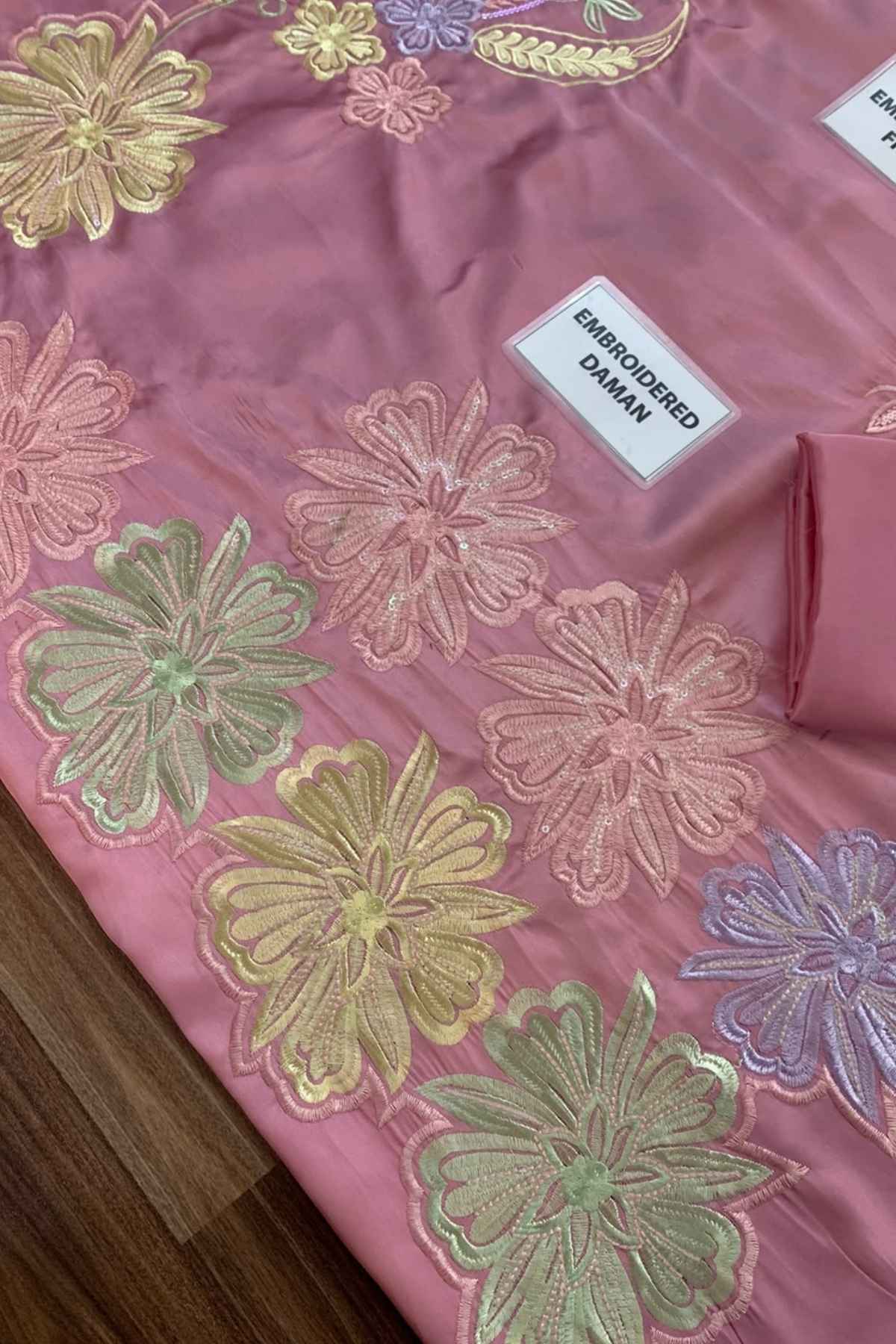 A close-up of a pink embroidered fabric, likely a saree or textile, featuring large floral motifs in gold, green, and purple thread. A white label reading 'EMBROIDERED DAMAN' is stitched onto the fabric, which is laid over a dark wood floor.