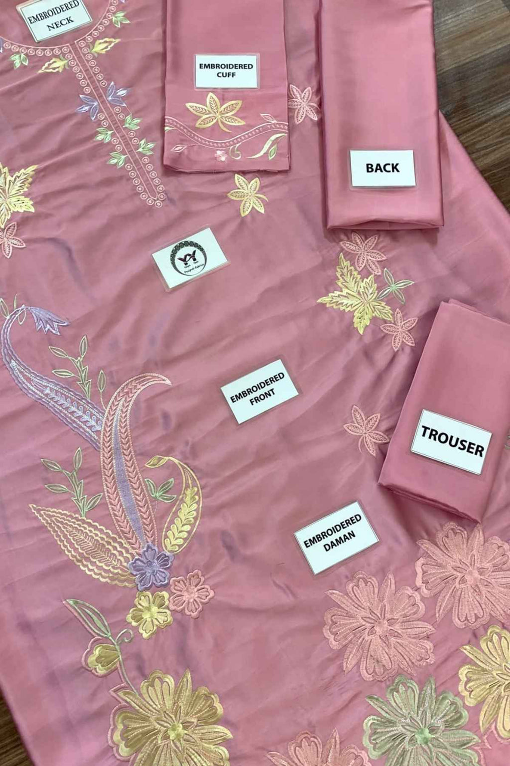 A dusty pink embroidered fabric set laid out on a wooden surface, including a kurta piece, matching pants, and accessories with labels pointing to embroidered sections.