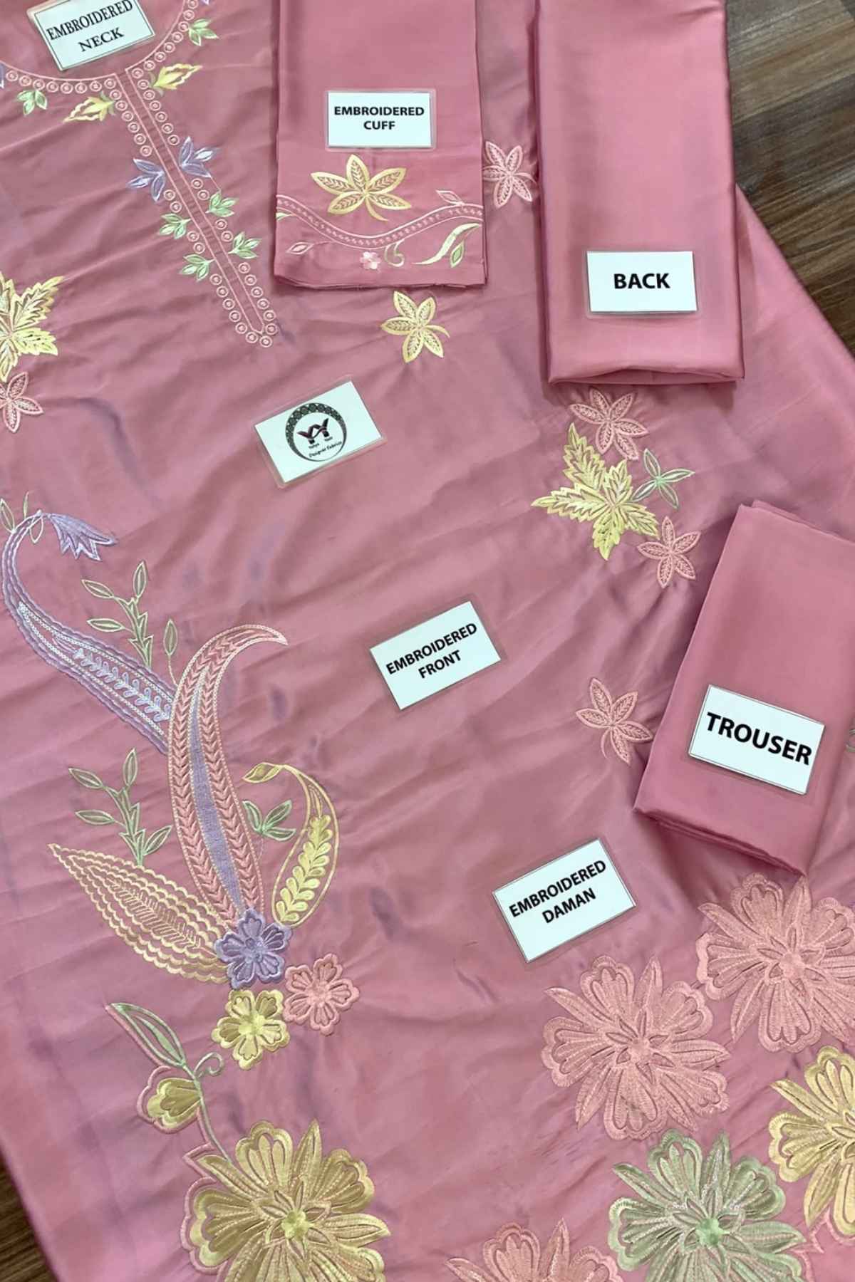 A dusty pink embroidered fabric set laid out on a wooden surface, including a kurta piece, matching pants, and accessories with labels pointing to embroidered sections.
