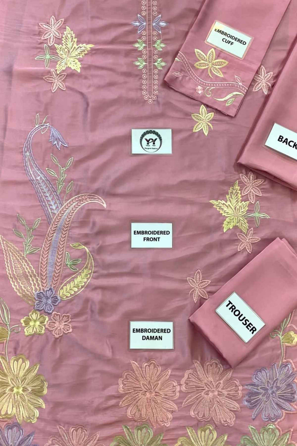 A dusty pink embroidered fabric set laid flat, including a kurta front piece, trouser piece, and matching fabric pieces labeled 'EMBROIDERED CURT', 'EMBROIDERED DAMAN', and 'BACK'. The fabric features multicolor floral and paisley embroidery in yellow, green, and lavender tones.