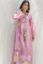A woman modeling a long pink embroidered kaftan dress with colorful floral and paisley patterns, standing against a white draped fabric background.