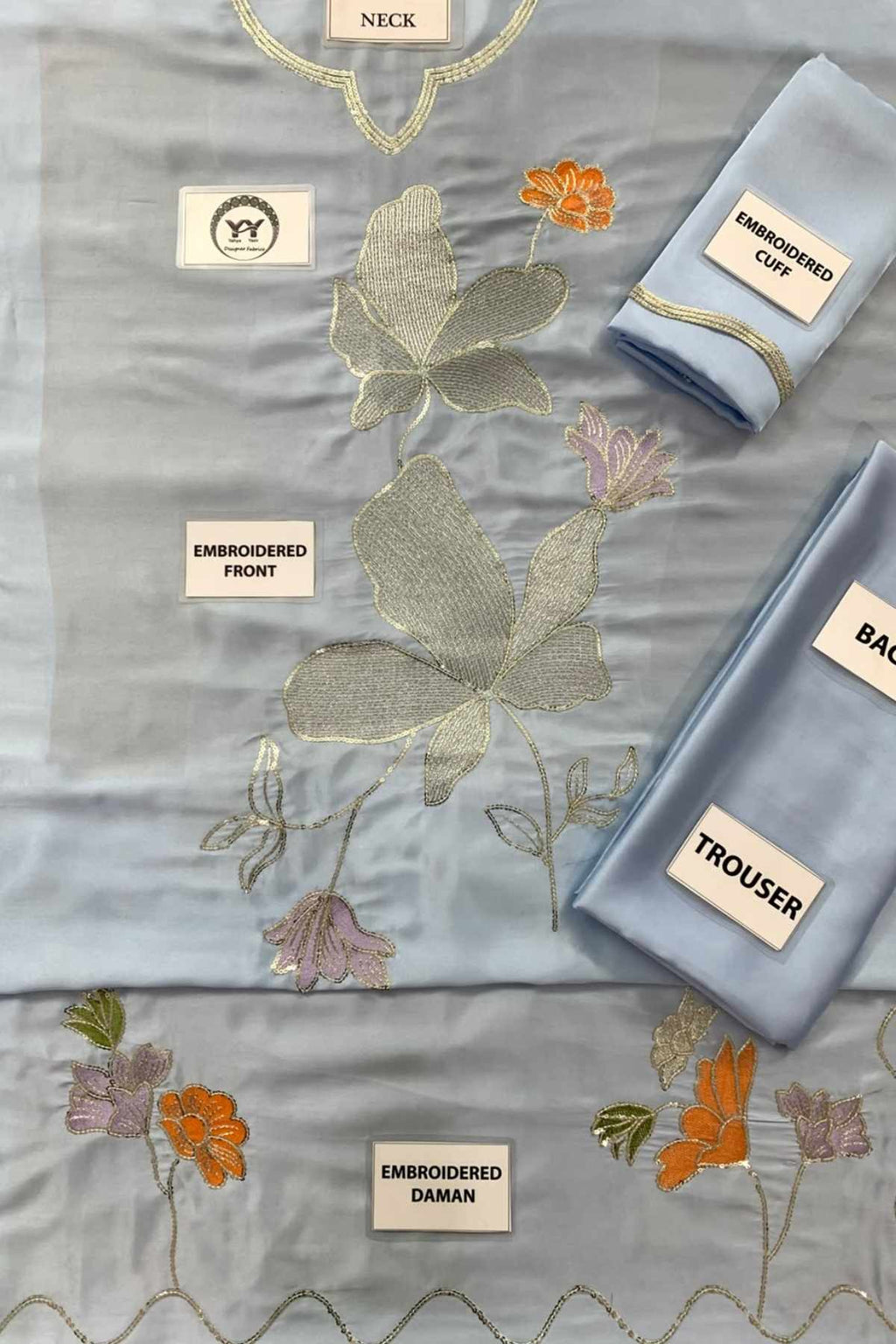 A light blue embroidered fabric set laid out on a white background, including a main embroidered piece labeled 'NECK', a folded piece labeled 'EMBROIDERED OFF', a folded piece labeled 'BACK', and a folded piece labeled 'TROUSER'. The fabric features metallic gold embroidery, peach-orange floral motifs, and lavender-pink floral accents.