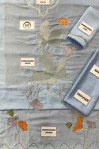 A light blue embroidered fabric set laid out on a white background, including a main embroidered piece labeled 'NECK', a folded piece labeled 'EMBROIDERED OFF', a folded piece labeled 'BACK', and a folded piece labeled 'TROUSER'. The fabric features metallic gold embroidery, peach-orange floral motifs, and lavender-pink floral accents.