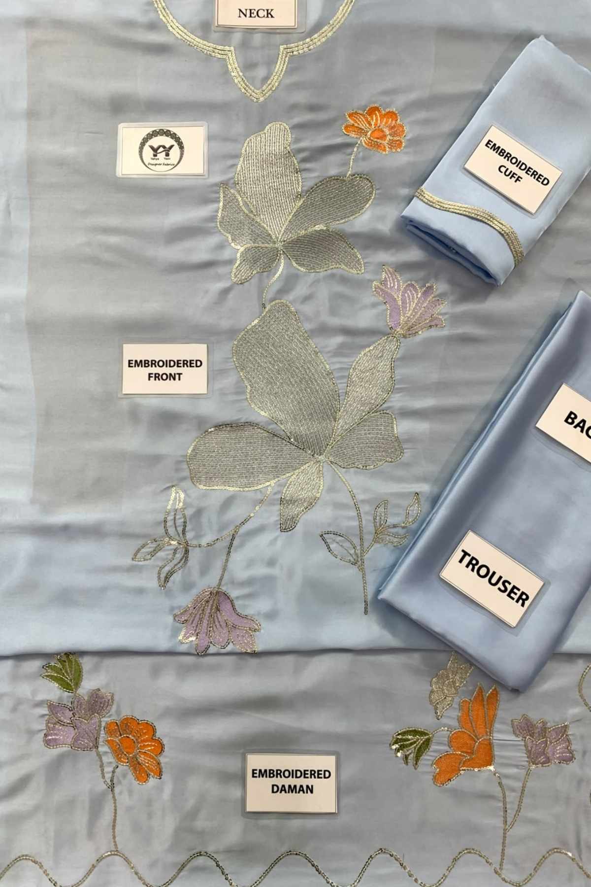 A light blue embroidered fabric set laid out on a white background, including a main embroidered piece labeled 'NECK', a folded piece labeled 'EMBROIDERED OFF', a folded piece labeled 'BACK', and a folded piece labeled 'TROUSER'. The fabric features metallic gold embroidery, peach-orange floral motifs, and lavender-pink floral accents.