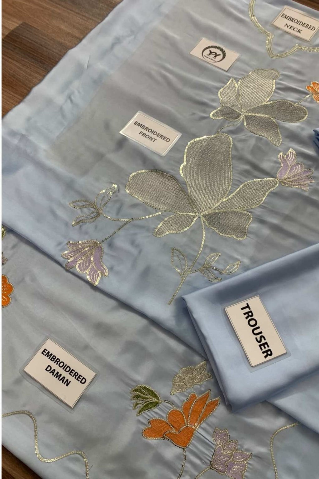 A folded light blue fabric garment set with embroidered floral motifs in gold, orange, and pink, shown on a wooden surface with fabric labels visible.