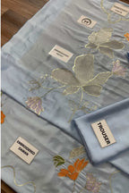 A folded light blue fabric garment set with embroidered floral motifs in gold, orange, and pink, shown on a wooden surface with fabric labels visible.