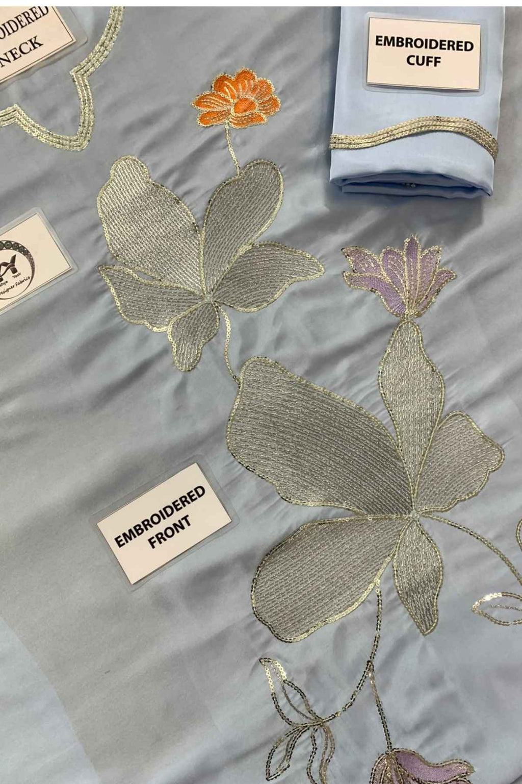 A light blue embroidered fabric garment laid flat, showing large metallic silver and gold floral motifs, an orange flower, and a folded matching cuff with gold trim. White labels identify parts such as the embroidered cuff, embroidered front, and embroidered neck.