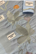 A light blue embroidered fabric garment laid flat, showing large metallic silver and gold floral motifs, an orange flower, and a folded matching cuff with gold trim. White labels identify parts such as the embroidered cuff, embroidered front, and embroidered neck.