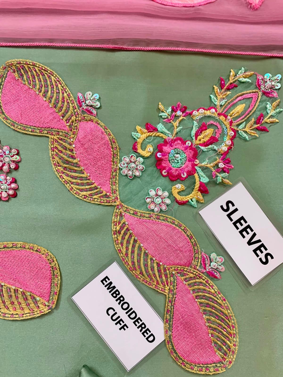 A styled flat-lay of a pink and mint green embroidered traditional outfit fabric set, showing various labeled components like the dupatta, front, trooper, and neck on a wooden surface.