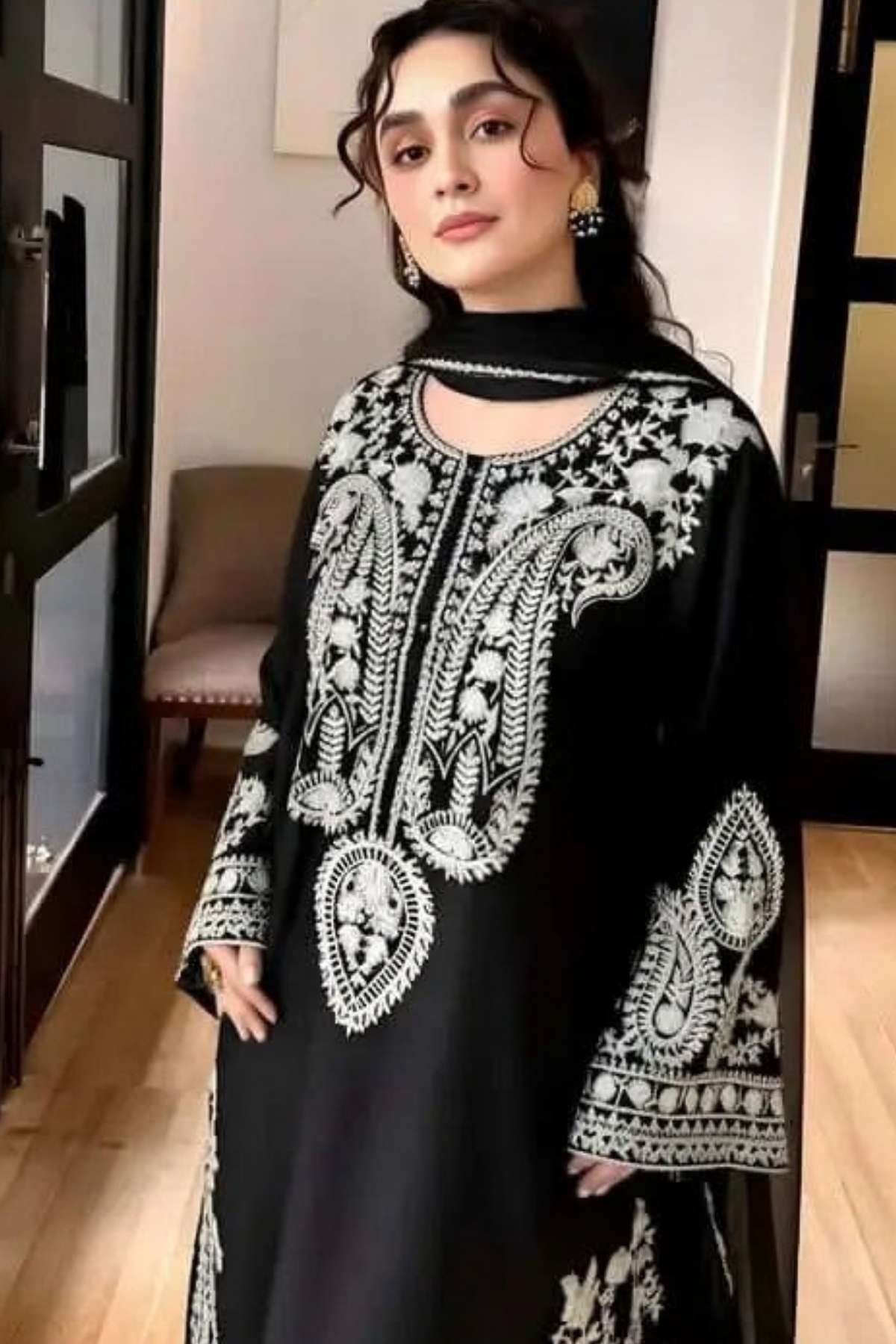 A woman wearing a black long kurta with intricate white embroidery and a matching black scarf, standing in an indoor hallway.