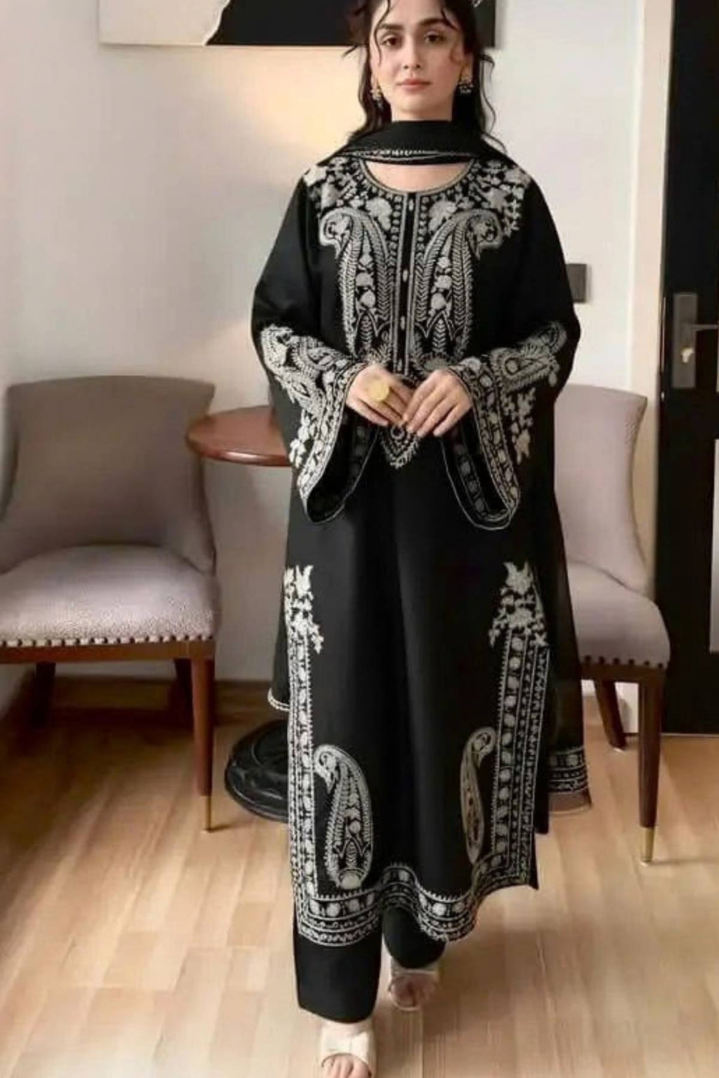 A woman wearing a black long kurta with intricate white embroidery, standing in a modern indoor room with light wood floors and gray upholstered chairs.