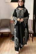 A woman wearing a black long kurta with intricate white embroidery, standing in a modern indoor room with light wood floors and gray upholstered chairs.