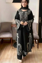 A woman wearing a black long kurta with intricate white embroidery, standing in a modern indoor room with light wood floors and gray upholstered chairs.