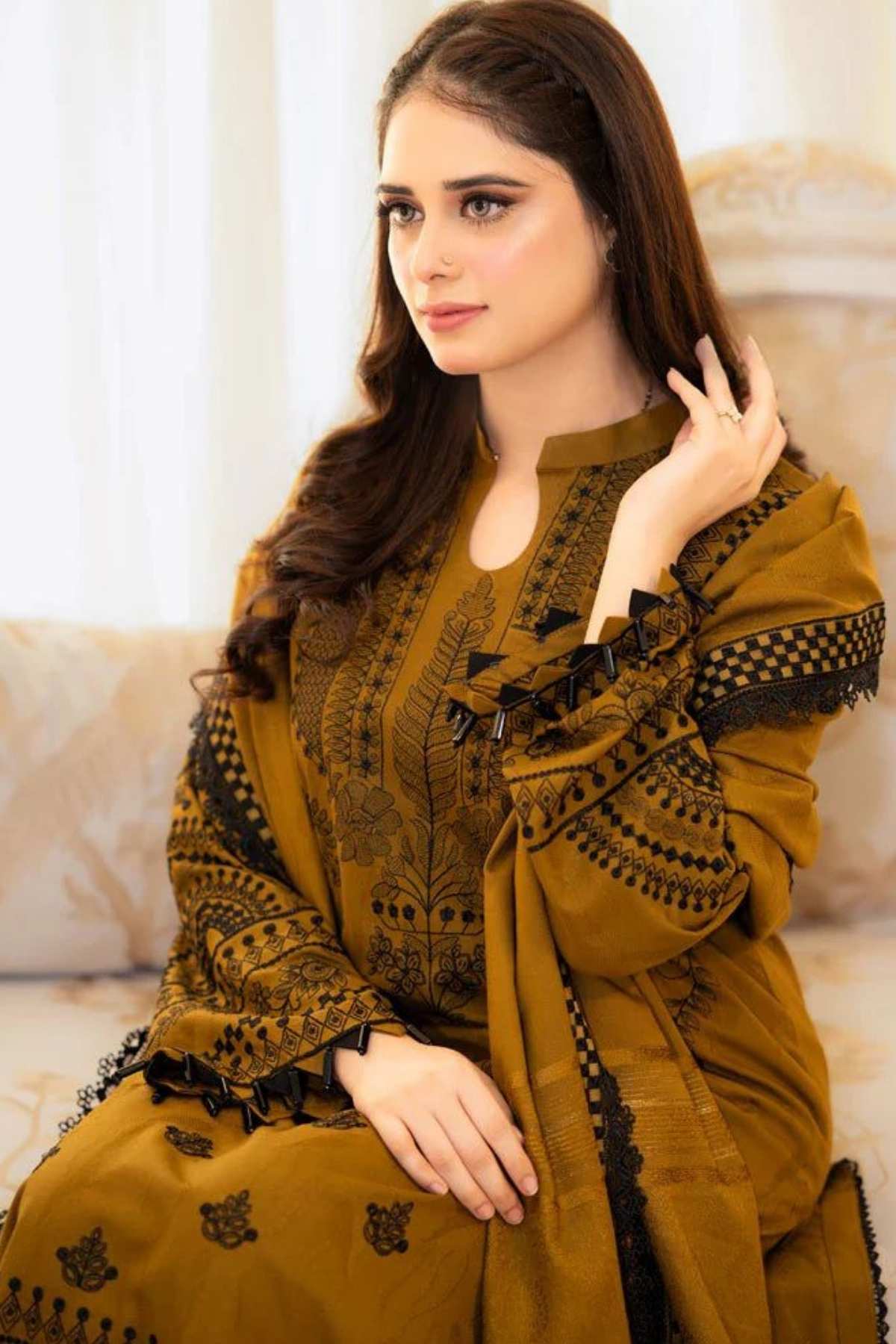 A woman wearing a mustard yellow embroidered traditional outfit with black detailing, seated on a light-colored sofa in an indoor setting.