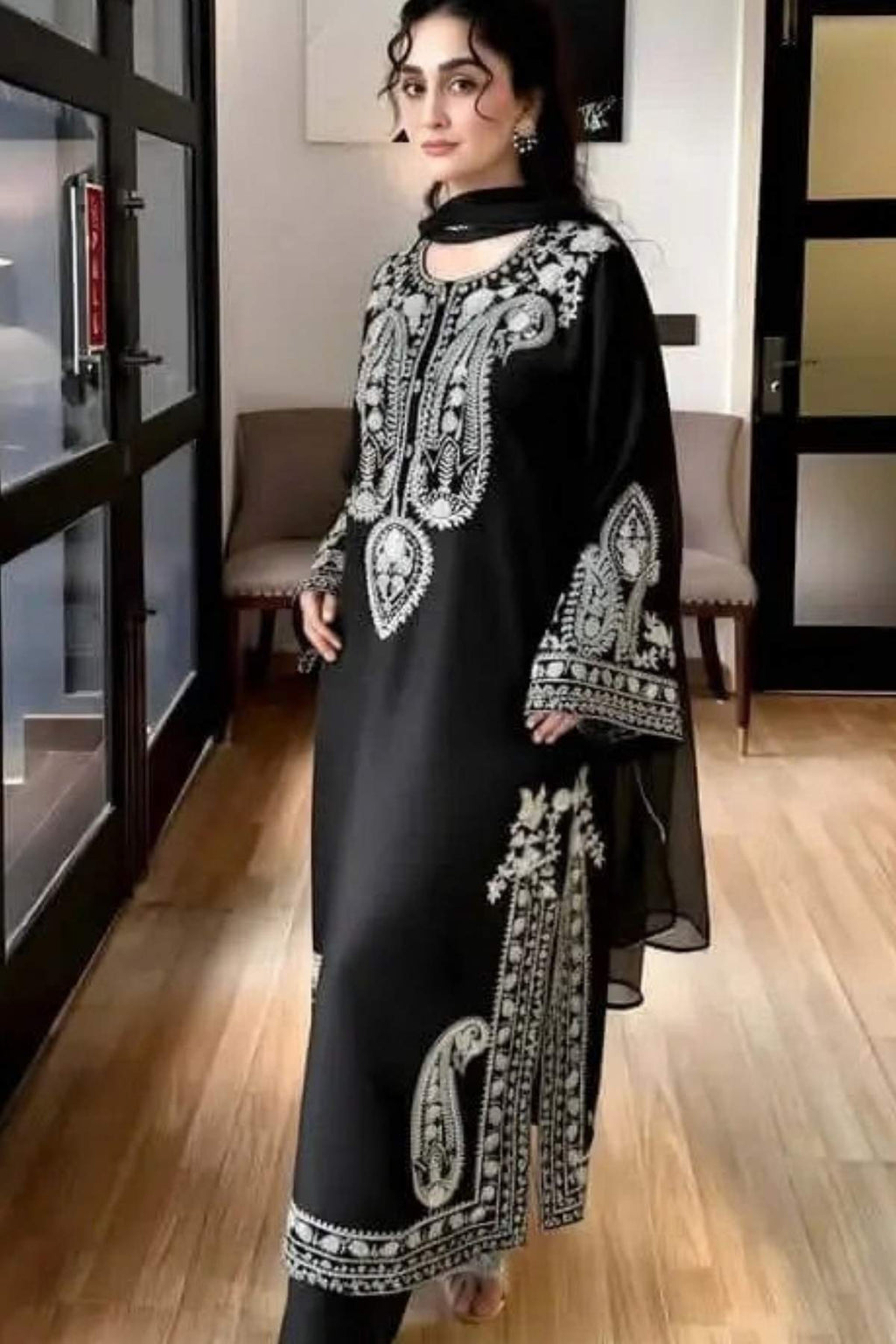 A woman modeling a black traditional outfit with intricate silver and white embroidery, standing in an indoor setting with wooden floors and glass doors.