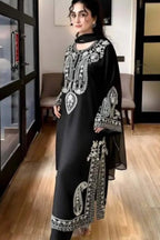 A woman modeling a black traditional outfit with intricate silver and white embroidery, standing in an indoor setting with wooden floors and glass doors.