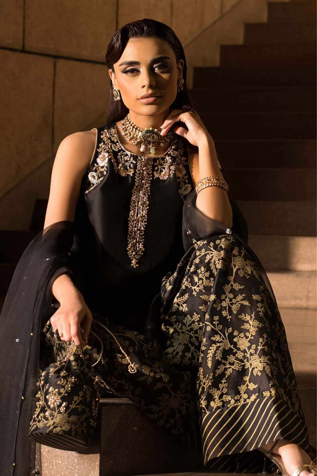 A woman seated on stone stairs wearing an ornate black and gold embroidered traditional outfit with a matching dupatta, heavy gold jewelry, and a matching handbag.