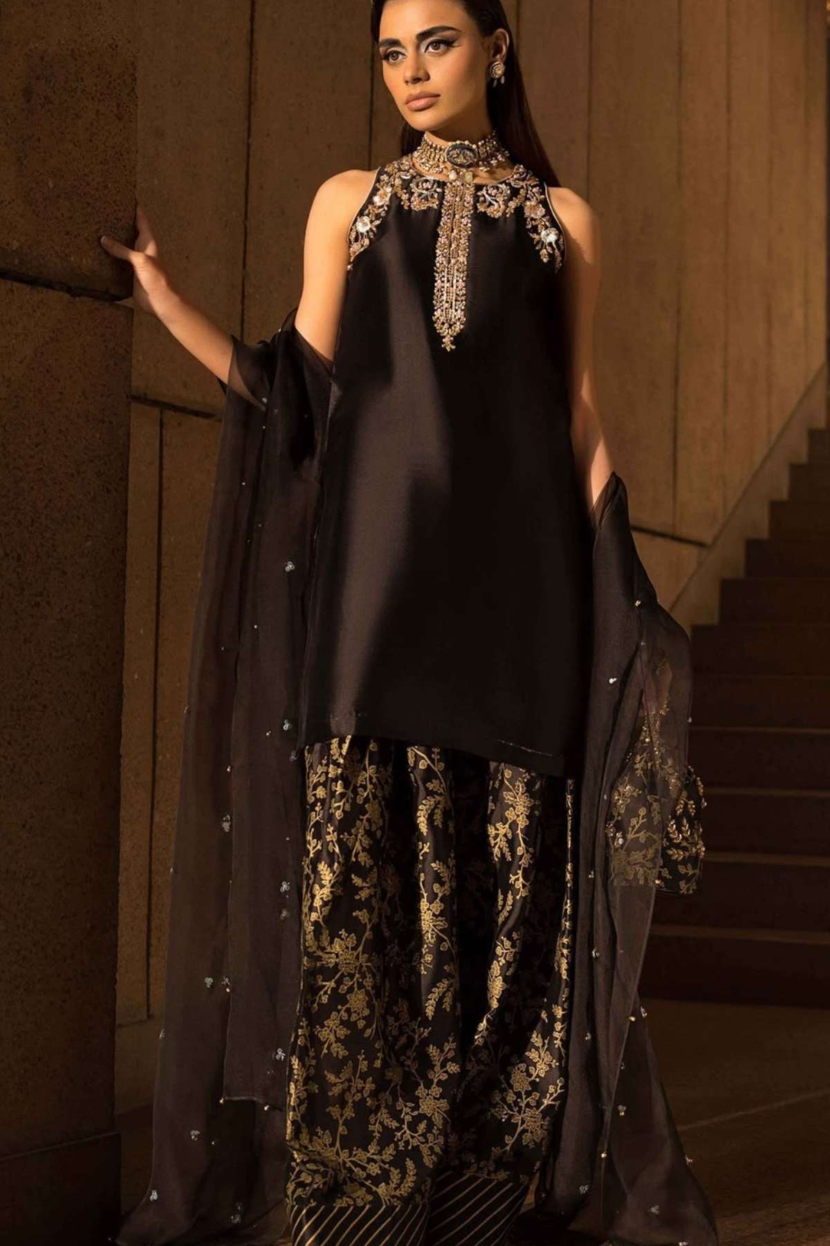 A woman models a black three-piece traditional outfit with gold embroidery and a sheer black dupatta in an indoor setting with warm lighting.