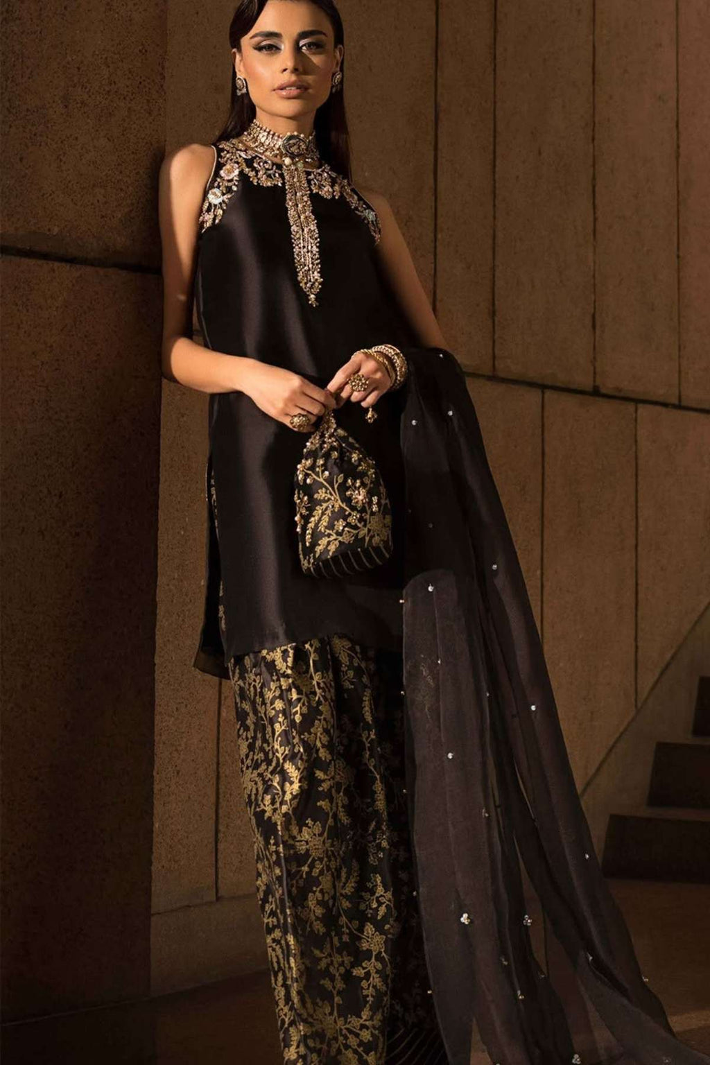 A woman models a black three-piece traditional outfit with gold embroidery and a sheer black dupatta in an indoor setting with warm lighting.