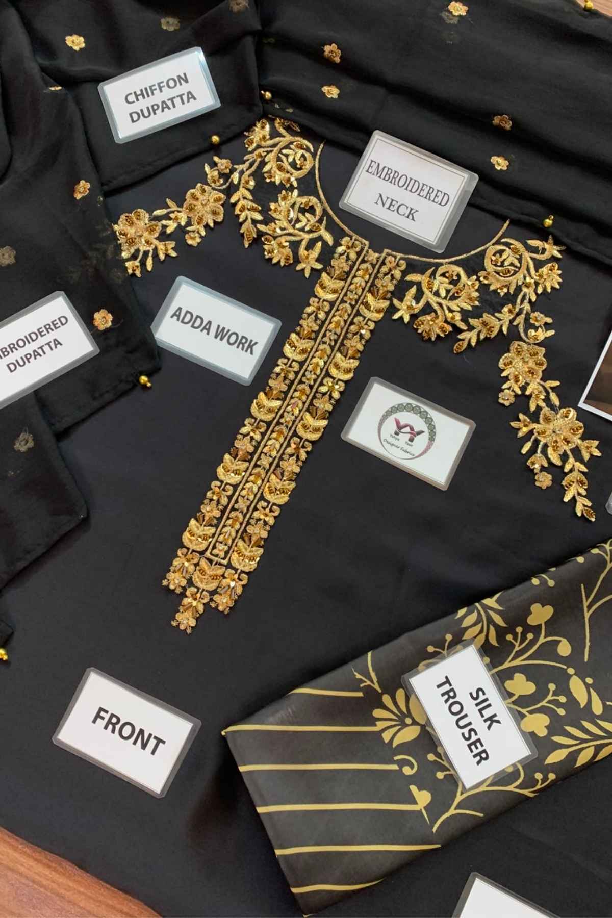 Black embroidered kurta set with gold floral embroidery, labeled parts including chevron dupatta, embroidered neck, adda work, front, and silk trouser, arranged on a dark surface.