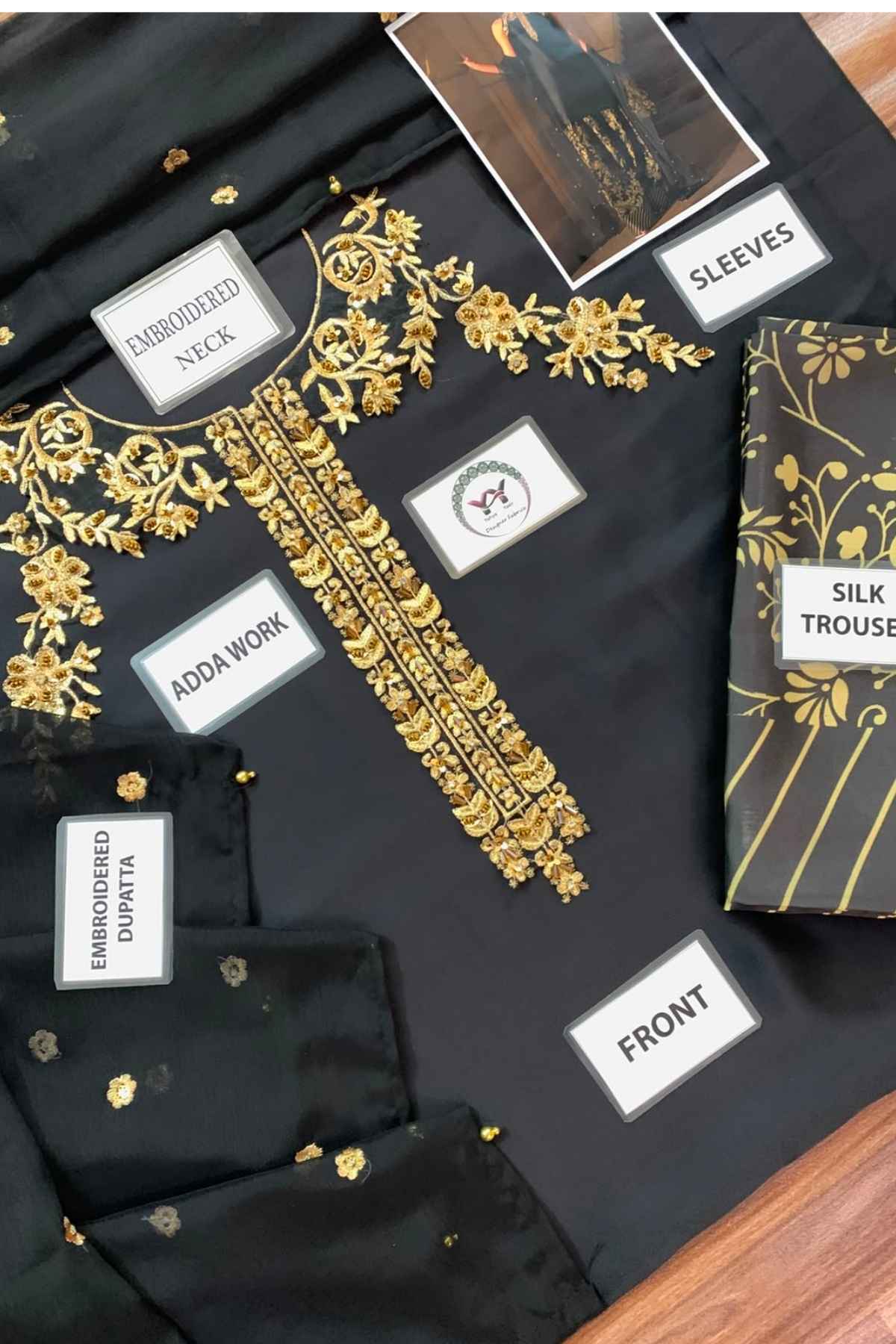 Black silk kurta set with intricate gold embroidery, shown with labeled fabric pieces including the front, sleeves, embroidered neck, and adda work on a wooden surface.