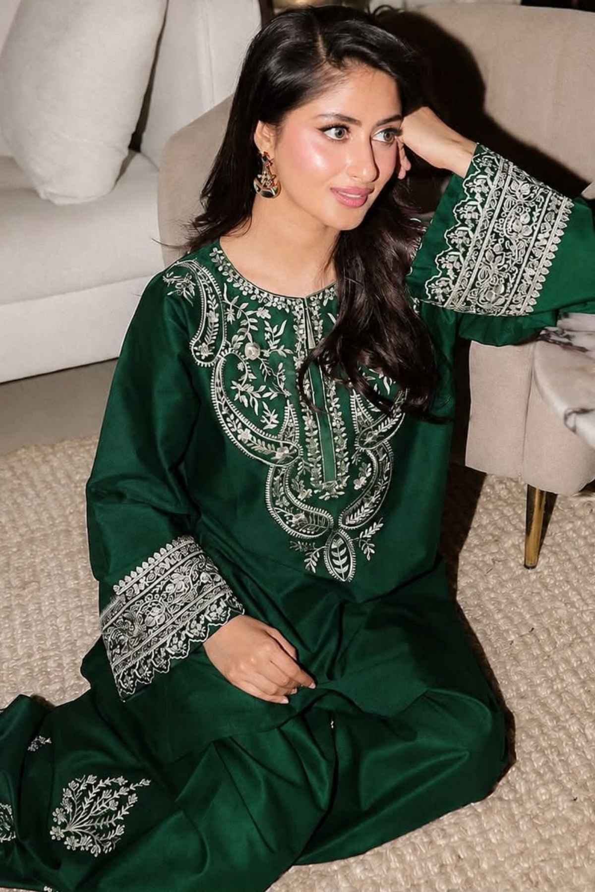 A woman wearing a dark green traditional outfit with intricate silver embroidery, sitting on a textured beige carpet in a living room setting.