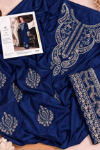 A royal blue fabric garment laid out on a beige surface, featuring ornate silver and light pink floral embroidery. A printed fashion card is placed on top of the fabric.