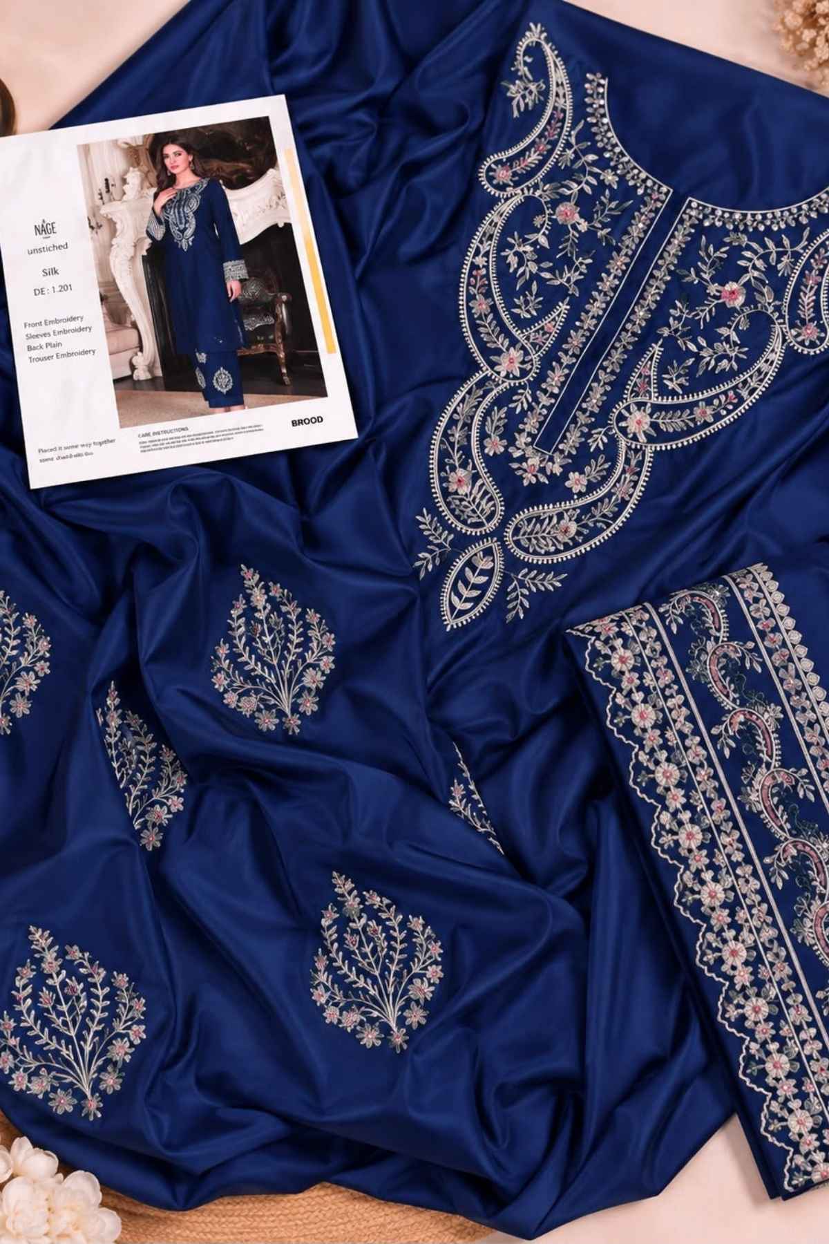 A royal blue fabric garment laid out on a beige surface, featuring ornate silver and light pink floral embroidery. A printed fashion card is placed on top of the fabric.