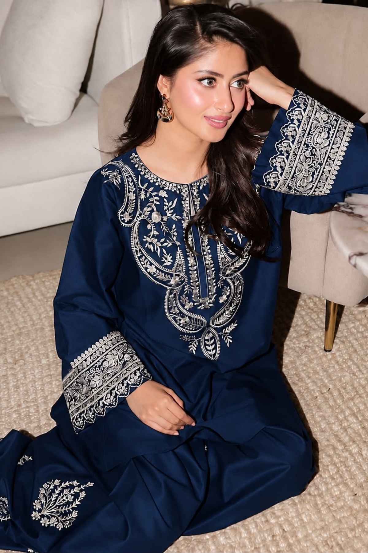 A woman wearing a navy blue traditional outfit with intricate silver embroidery, sitting on a textured beige rug in a modern living room setting.