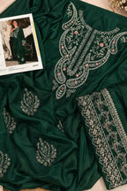 A dark green embroidered traditional outfit laid flat, featuring ornate silver and pink floral embroidery. A folded matching embroidered dupatta or fabric piece is placed beside it, and a small printed fashion reference card is visible in the upper left on a light beige surface.
