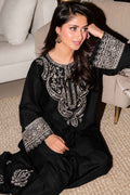 A woman wearing a black traditional embroidered outfit with intricate silver-white detailing on the chest and cuffs, sitting on a textured beige rug in a modern living room setting.