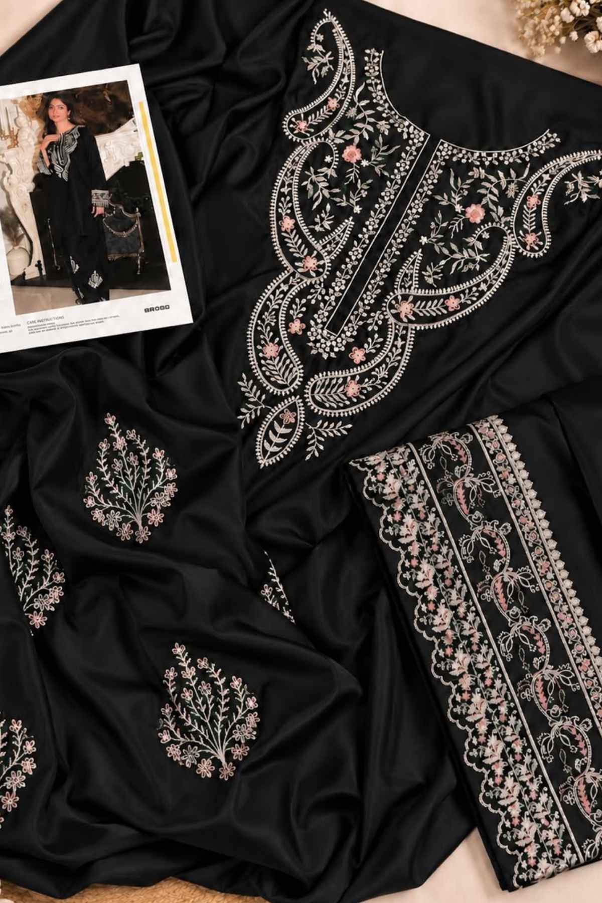 Black fabric garment laid flat with ornate silver and pink floral embroidery, shown with matching embroidered border detail and a small printed fashion insert on a neutral surface.
