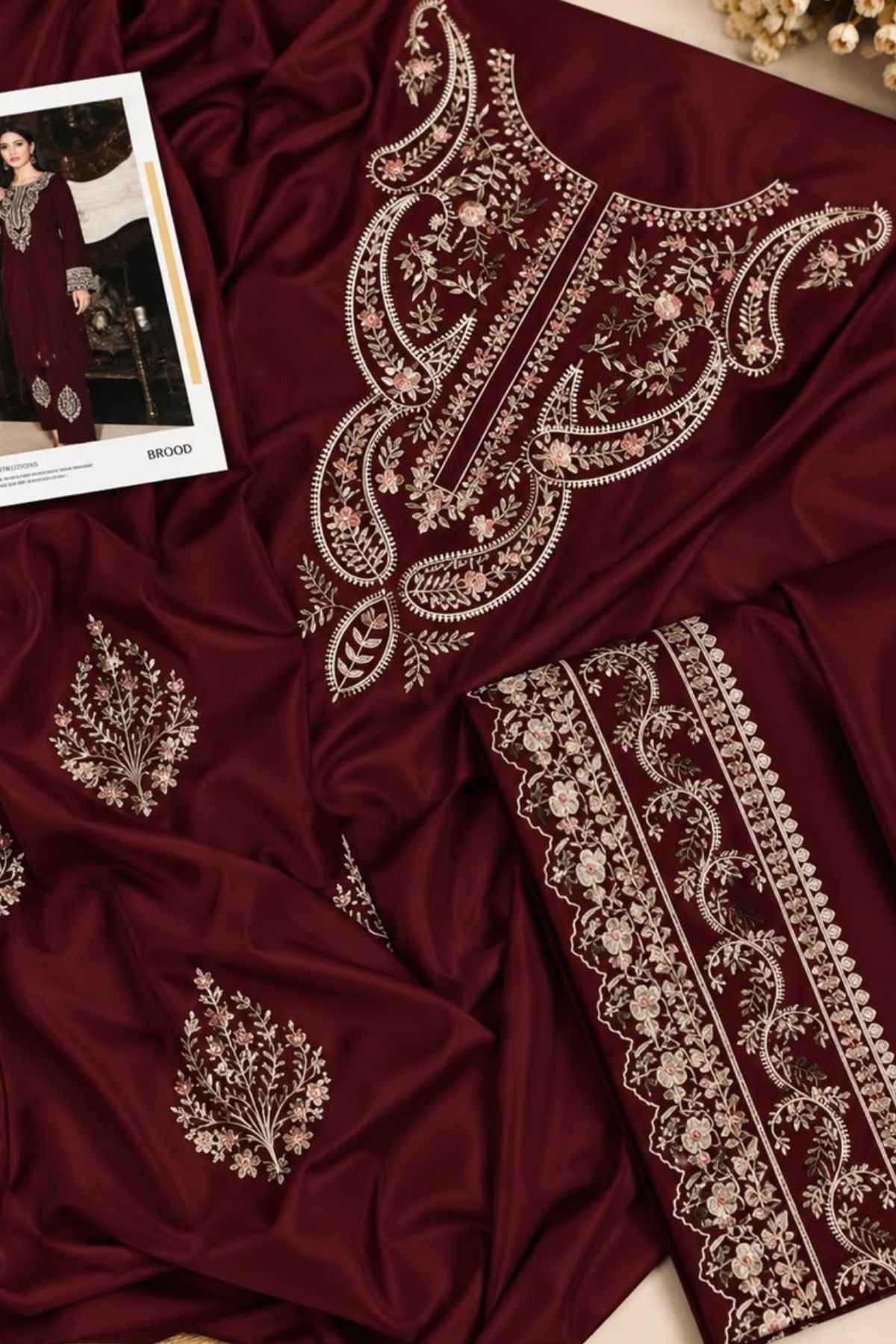 A deep maroon/burgundy fabric set with ornate beige and gold floral embroidery, shown in a styled flat-lay arrangement with a small printed fashion card in the top-left corner.